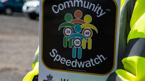 A person in a high visibility jacket holds a clipboard which says Community Speedwatch